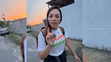 A candidata Kelaine Goulart caminha por uma rua de Itaguaí, olhando para a câmera e apontando o dedo indicador para cima enquanto fala. Ela veste uma camiseta branca de campanha e mochila nas costas, cercada por moradores e apoiadores ao ar livre no fim de tarde.