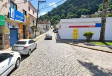 Rua pavimentada no Centro de Mangaratiba, com veículos, lojas, rede elétrica e morro arborizado ao fundo.