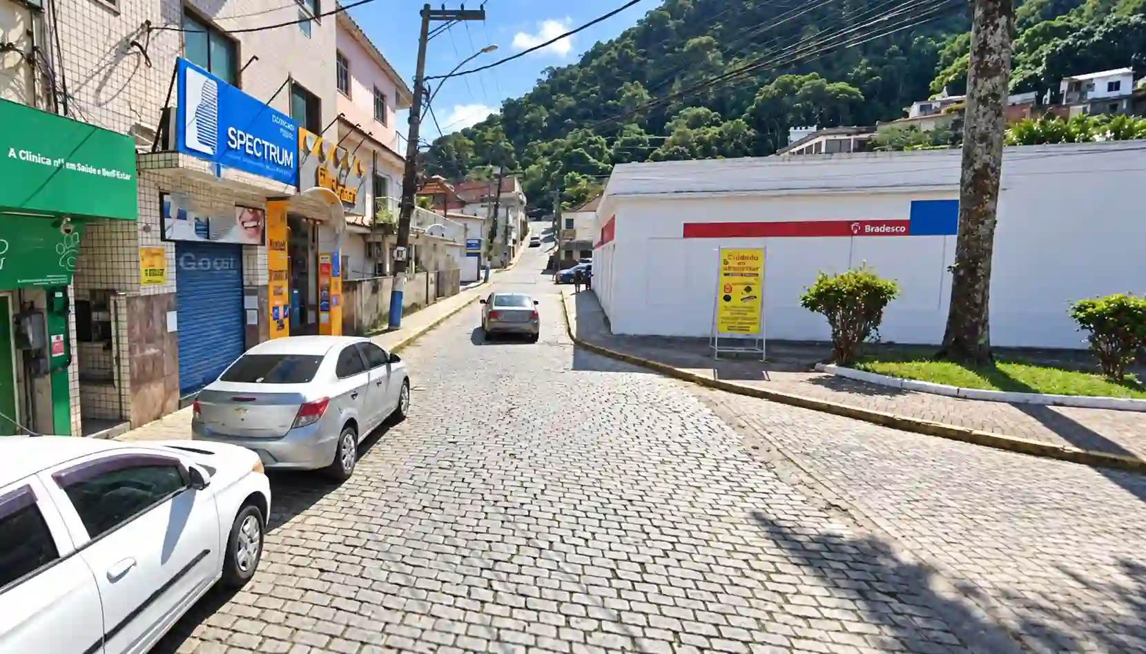 Rua pavimentada no Centro de Mangaratiba, com veículos, lojas, rede elétrica e morro arborizado ao fundo.