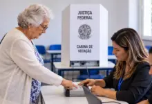 Na foto gerada por IA, Mulher idosa realiza biometria em seção eleitoral, diante da mesa receptora com duas mesárias, com cabine de votação ao fundo.