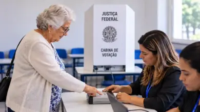 Na foto gerada por IA, Mulher idosa realiza biometria em seção eleitoral, diante da mesa receptora com duas mesárias, com cabine de votação ao fundo.