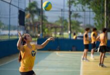 Menina com camiseta amarela do VivaVôlei prepara um saque em quadra ao ar livre, com a bola lançada acima da cabeça e um braço levantado. Outros alunos aparecem ao fundo.