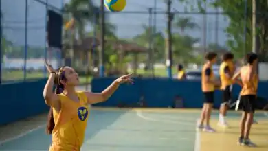 Menina com camiseta amarela do VivaVôlei prepara um saque em quadra ao ar livre, com a bola lançada acima da cabeça e um braço levantado. Outros alunos aparecem ao fundo.