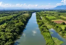 Vista aérea do Rio Guandu, cercado por áreas de vegetação, campos e linhas de transmissão de energia, com montanhas ao fundo.