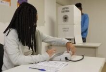 Mesária opera equipamento de identificação do eleitor em uma seção eleitoral, enquanto uma pessoa está na cabine de votação ao fundo.