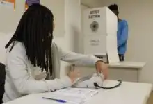 Mesária opera equipamento de identificação do eleitor em uma seção eleitoral, enquanto uma pessoa está na cabine de votação ao fundo.
