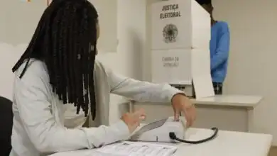 Mesária opera equipamento de identificação do eleitor em uma seção eleitoral, enquanto uma pessoa está na cabine de votação ao fundo.
