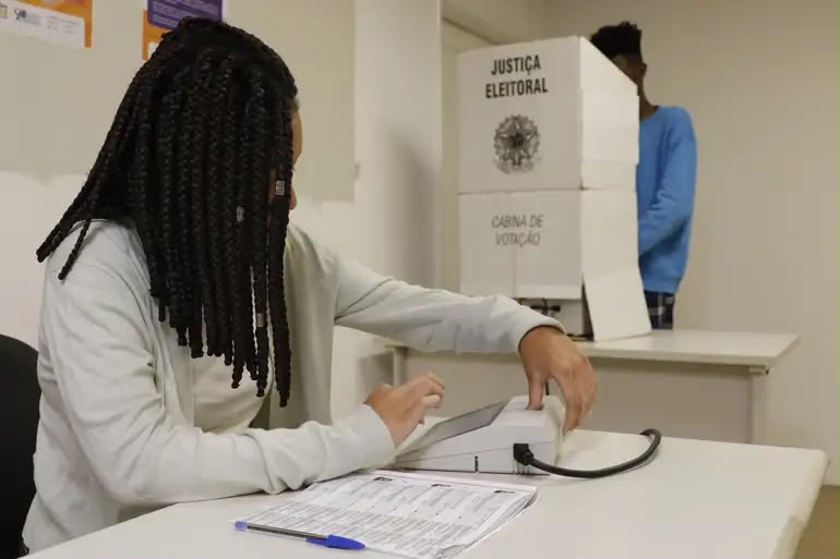 Mesária opera equipamento de identificação do eleitor em uma seção eleitoral, enquanto uma pessoa está na cabine de votação ao fundo.