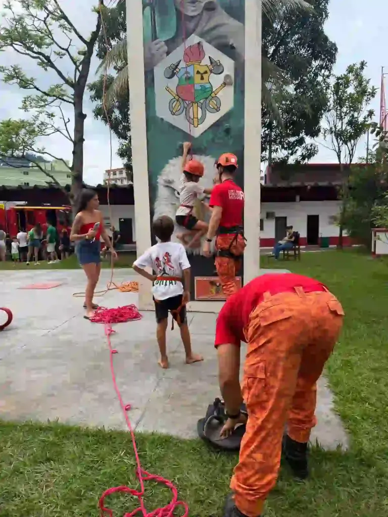 Criança com capacete e equipamento de segurança participa de atividade de escalada acompanhada por um bombeiro, enquanto outra aguarda sua vez no quartel.